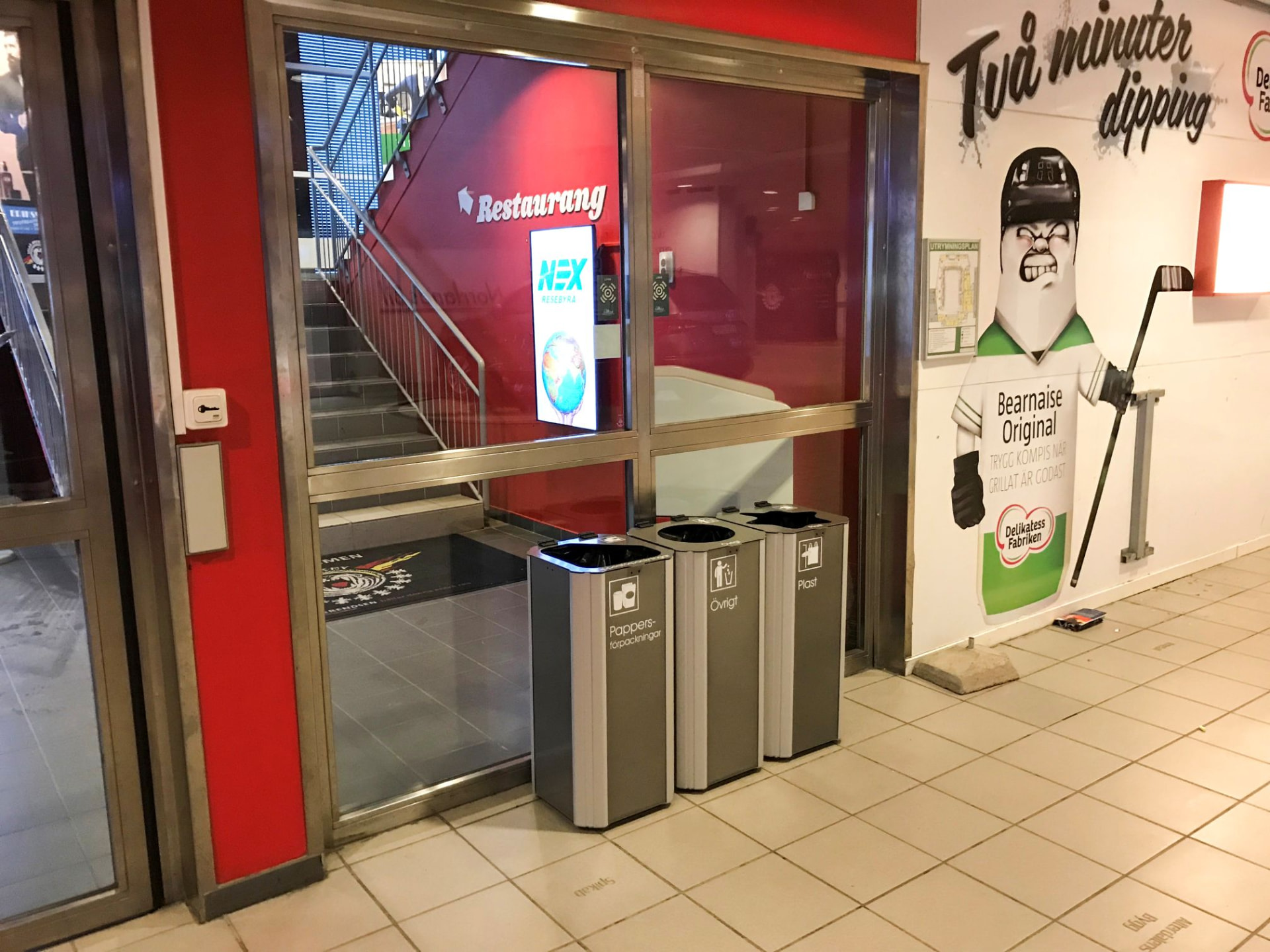 Tre grå Kiwi kärl med märkning för pappersförpackningar, övrigt och plast vid restaurangentrén i en arena