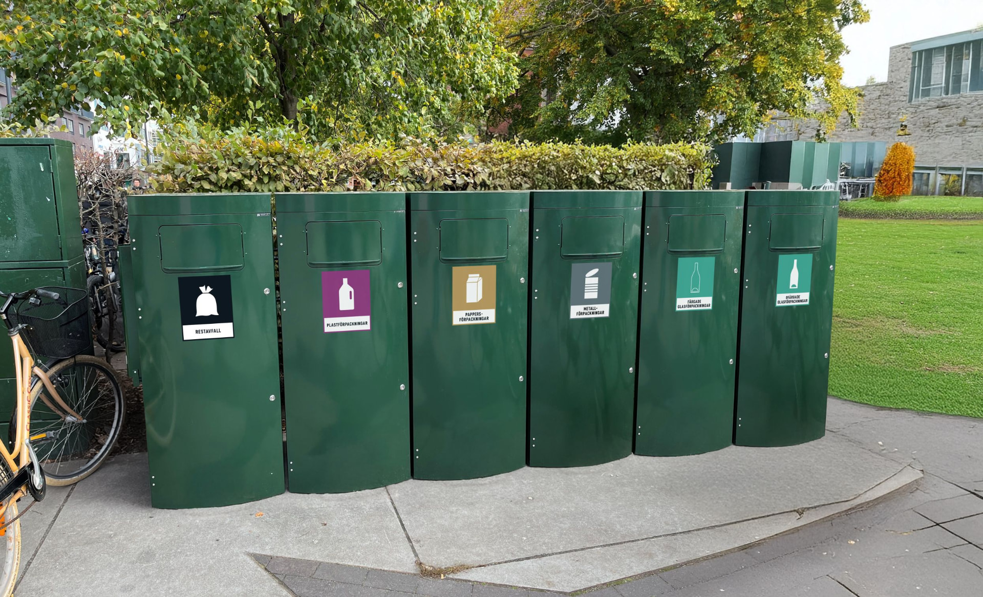Sex gröna Sortify Mango kärl för sex fraktioner vid en häck intill en gräsyta och cykelparkering