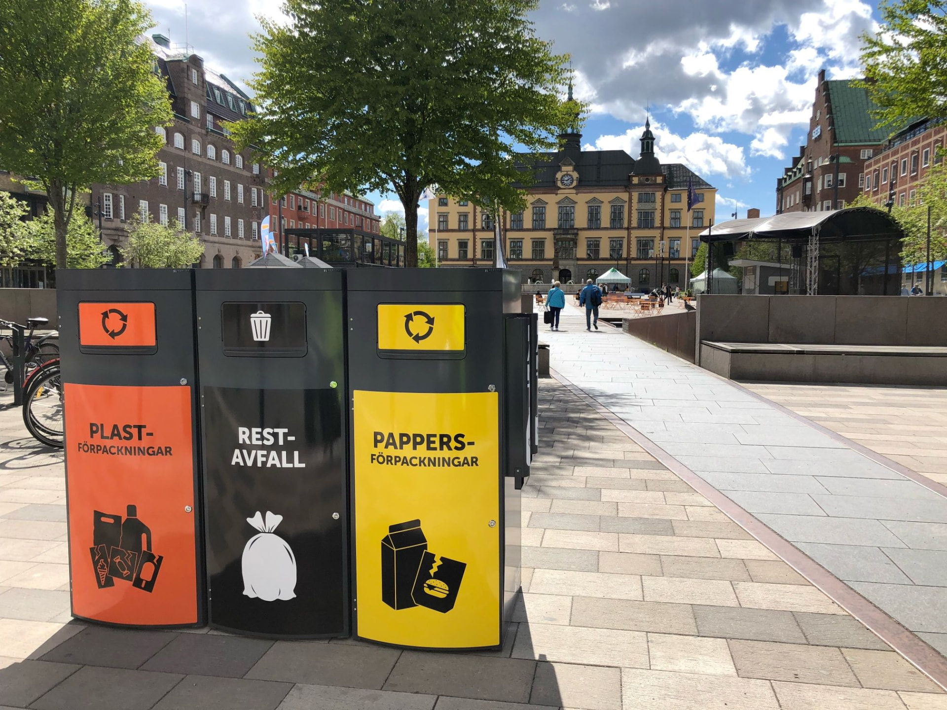 Tre Sortify Mango kärl i orange, svart och gult för plast, restavfall och papper på ett torg i Eskilstuna