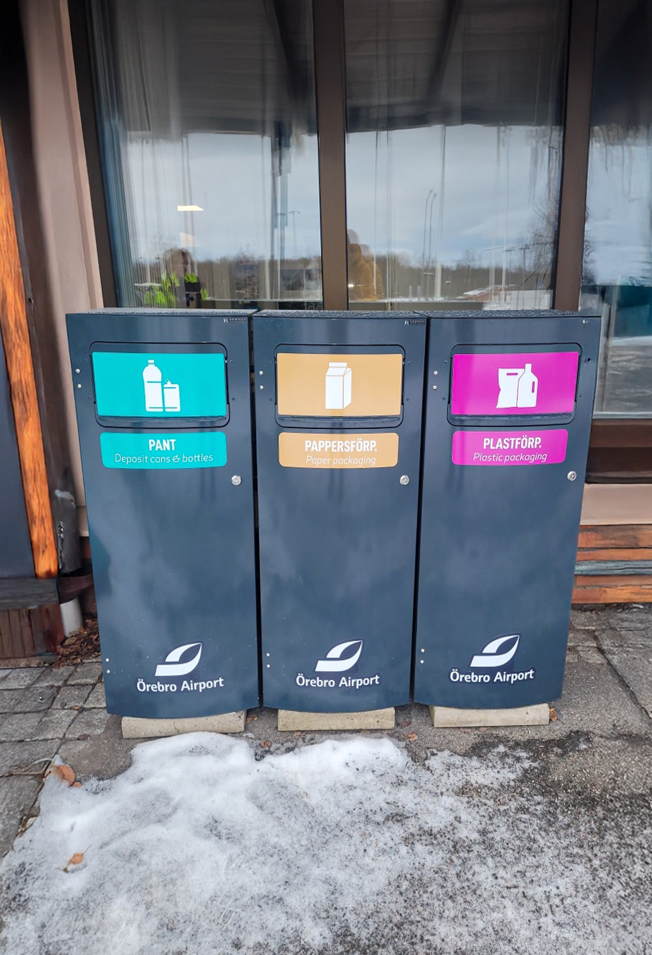 Tre mörka Mango M kärl för pant, pappersförpackningar och plastförpackningar med Örebro Airports logotyp vid en entré