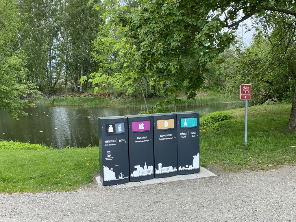 Fyra mörka Mango M kärl med Vänersborgs kommuns stadssiluett vid en parkdamm