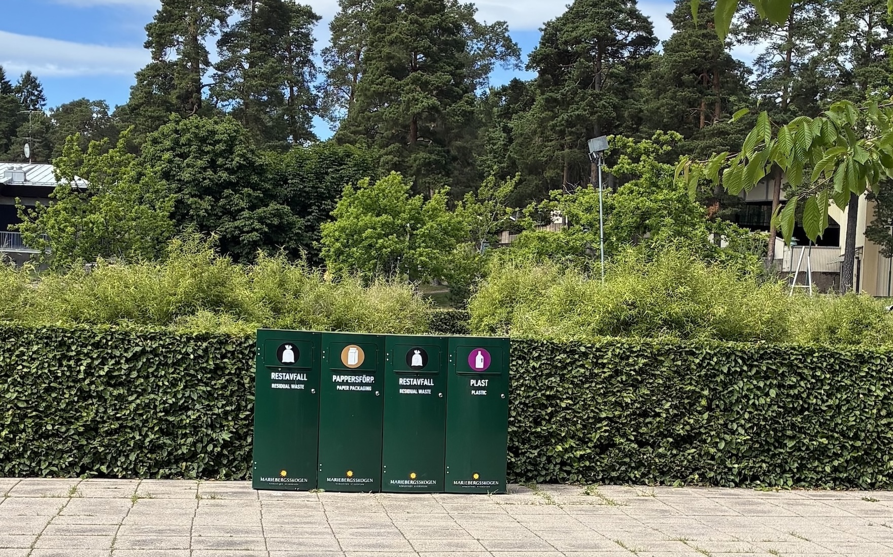 Fyra mörkgröna Mango M kärl för restavfall, pappersförpackningar och plast med Mariebergsskogens tryck framför en klippt häck