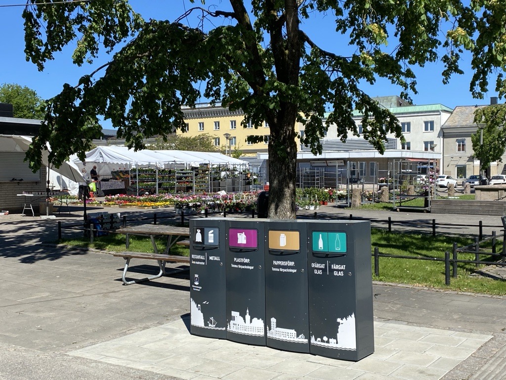 Fyra mörka Mango M avfallskärl för restavfall och metall, plastförpackningar, pappersförpackningar samt ofärgat och färgat glas vid ett torg i Vänersborg
